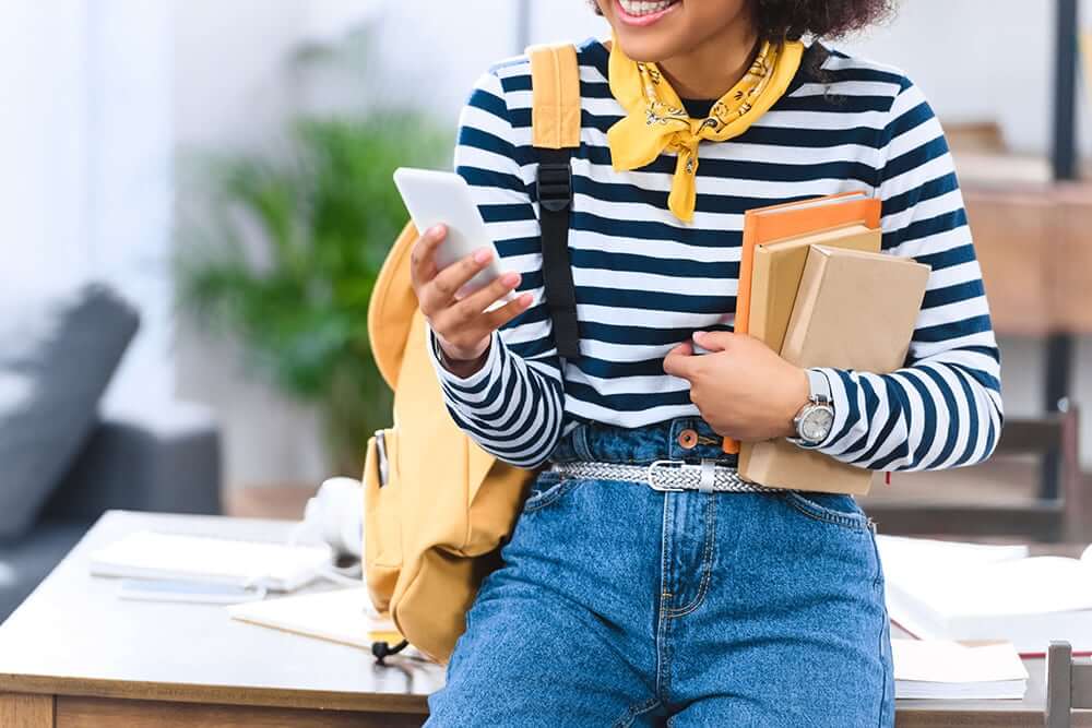 Woman sitting on desk holding books and looking at phone